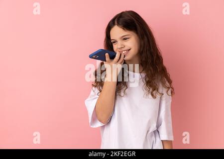 Tecnologia vocale intelligente mobile. Ritratto di una bambina che indossa una T-shirt bianca che parla con lo smartphone utilizzando l'assistente virtuale, l'app per altoparlanti digitali. Studio in interni isolato su sfondo rosa. Foto Stock