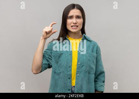 Piccola quantità. Ritratto di adorabile bella giovane donna che mostra un po 'di gesto e guardare pleadingly, indossando giacca casual stile. Studio in interni isolato su sfondo grigio. Foto Stock