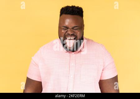 Ritratto dell'uomo che indossa una camicia rosa che sporge dalla lingua e guarda la fotocamera, brulicante di espressione naughty, comportamento disobbediente. Studio al coperto isolato su sfondo giallo. Foto Stock
