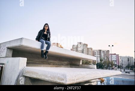 latina ragazza in pantaloni blu strappato pelliccia occhiali giacca sorridente alla fotocamera sorridente seduta calma e rilassato sulla parete di cemento in città Foto Stock