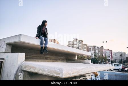 latina ragazza in pantaloni blu strappato pelliccia occhiali giacca guardando la città seduta calma e rilassato sulla parete di cemento Foto Stock