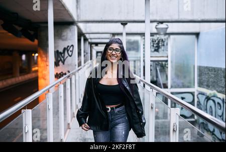 giovane latina donna occhiali bruna pelliccia giacca grande nero stivali con un grande sorriso felice e soddisfatto a piedi lungo il passaggio di vetro Foto Stock