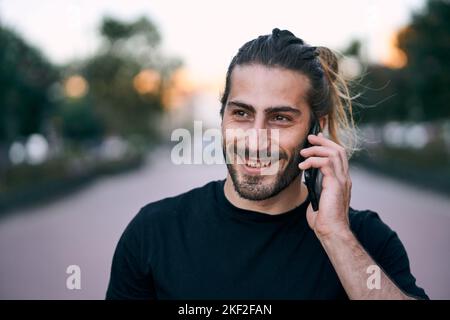 il ragazzo caucasico nero t-shirt disheveled capelli lunghi con una barba sorridente in piedi sulla strada parlando sul suo smartphone sul viale della città Foto Stock