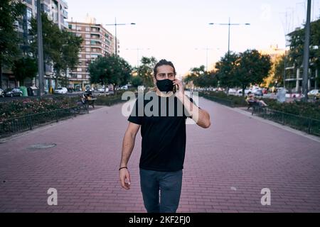 ragazzo caucasico dai capelli lunghi che cammina lungo il viale sorridendo parlando sullo smartphone guardando la macchina fotografica con la maschera sul suo volto in città Foto Stock