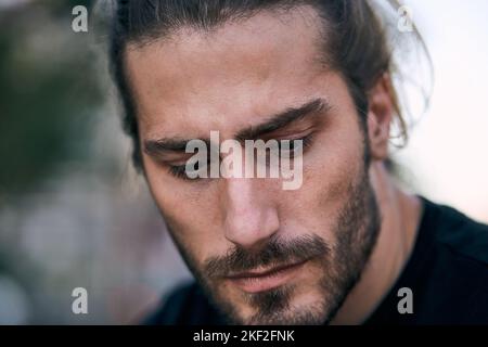 primo piano faccia serio caucasico ragazzo guardando giù con barba lunga capelli dispettati preoccupati infelici Foto Stock