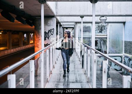 latina ragazza con lunghi capelli brunette occhiali pelliccia giacca pantaloni blu e grandi stivali neri labbro piercing a piedi sul passaggio di vetro Foto Stock