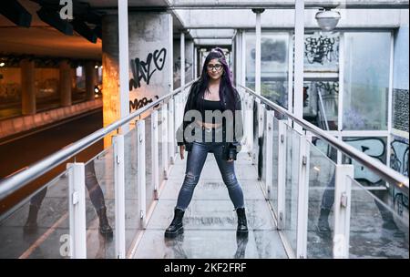 latina ragazza con lunghi capelli brunette occhiali pelliccia giacca strappato jeans blu e grandi stivali neri in piedi guardando la macchina fotografica con le gambe spalmata sul vetro Foto Stock