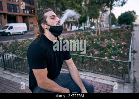 giovane uomo caucasico nero t-shirt seduta vaping espirando vapore acqueo con una maschera nera sotto la bocca in città Foto Stock