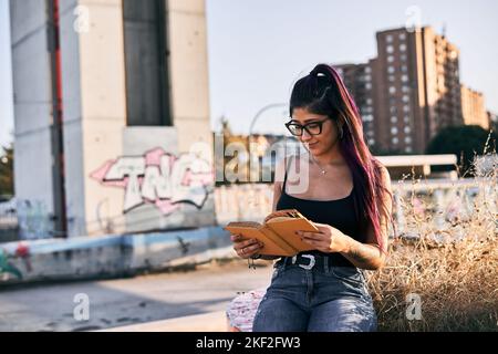 latina ragazza con occhiali brunette lunga collana capelli sul collo piercing sul suo labbro sorridente mentre legge un libro nel parco della città Foto Stock