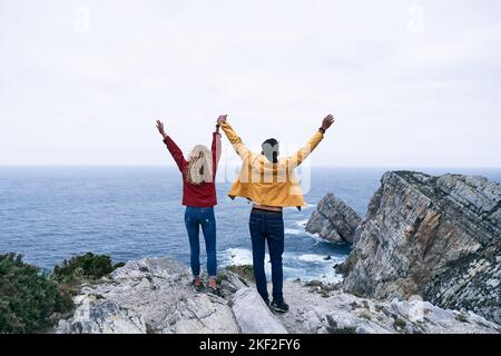 ragazzo latino e ragazza caucasica in giacca rossa e gialla pantaloni blu e nero in piedi tenendo le mani con le braccia tese dalle grandi rocce, cabo de Foto Stock