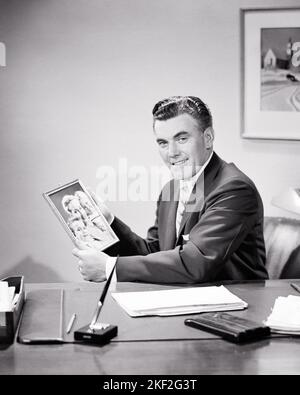 1950S RITRATTO SORRIDENTE UOMO D'AFFARI SEDUTO ALLA SUA SCRIVANIA CON UNA FOTO INCORNICIATA DI SUA MOGLIE E DEI SUOI FIGLI CHE GUARDANO LA MACCHINA FOTOGRAFICA - S11940 HAR001 HARS UOMINI D'AFFARI EXECUTIVE VECCHIO TEMPO NOSTALGIA VECCHIO MODO 1 GIOVANE STILE CARRIERA FAMIGLIE GIOIA LIFESTYLE SODDISFAZIONE FEMMINE LAVORO SPOSATO STUDIO SHOT CONIUGE MARITI SALUTE COPIA SPAZIO METÀ LUNGHEZZA PERSONE ISPIRAZIONE MASCHI CURA PROFESSIONE FIDUCIA DIRIGENTI B&W PARTNER OCCHIO CONTATTO SKILL SUIT E CRAVATTA PROFESSIONE FELICITÀ COMPETENZE SUO E CARRIERE BELLO ORGOGLIO OCCUPAZIONI BOSS CONNESSIONE INCORNICIATA ELEGANTE MEDIO-ADULTO MEDIO-ADULTO UOMO MOGLI DONNA DI METÀ-ADULTO Foto Stock