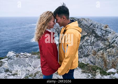 ragazzo latino e ragazza caucasica in giacca rossa e gialla di mezza lunghezza in piedi in profilo rivolto l'uno verso l'altro con la fronte in contatto sorridente tenuta Foto Stock