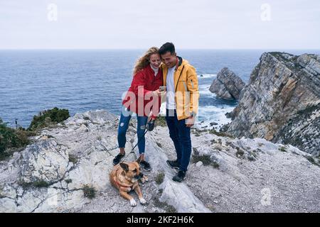 ragazza caucasica bionda in piedi sulle rocce vicino al mare accanto a un cane che mostra il suo smartphone schermo a un ragazzo latino con giacca gialla, cabo de penas Foto Stock