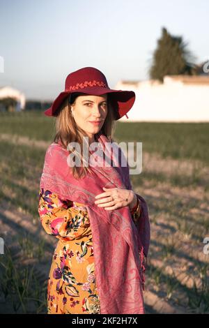 Donna matura caucasica in piedi in abito floreale e scialle rosa guardando la macchina fotografica sorridendo dal frutteto in una giornata di sole Foto Stock