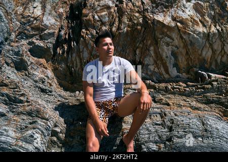 ragazzo latino dai capelli scuri con costume da bagno e t-shirt blu a piedi nudi seduto su una grande roccia calmo e rilassato in una giornata di sole, asturie, spagna Foto Stock