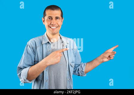 Studio shot di un uomo ispanico che indossa una camicia in denim in piedi su sfondo isolato blu sorridendo e guardando la macchina fotografica che punta con due mani e. Foto Stock
