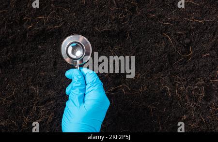 Mano di ricercatore che tiene uno stetoscopio su fertile suolo nero Foto Stock