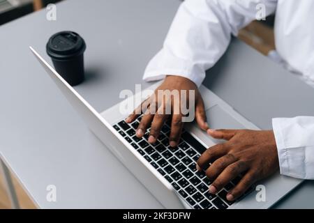 Primo piano ripresa ritagliata di un medico maschio afro-americano irriconoscibile in cappotto bianco che lavora digitando sul computer portatile seduto alla scrivania con caffè Foto Stock