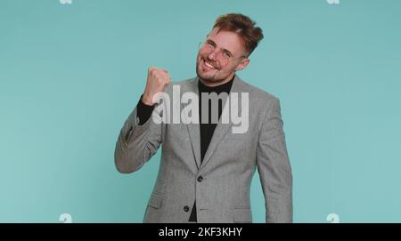 Felice uomo di ufficio gioioso di affari urlando, sollevando pugni nel gesto l'ho fatto, celebrando il successo, vincendo, compleanno, obiettivo della lotteria Achiemen. Ragazzo adulto al coperto su sfondo blu parete studio Foto Stock