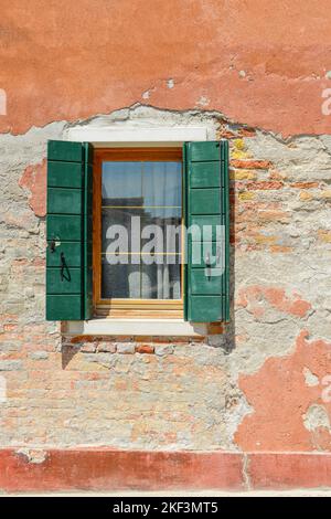 La finestra verde sul muro arancione della vecchia casa si rompe in mattoni rossi visibili in cemento a Burano, Italia. Foto Stock