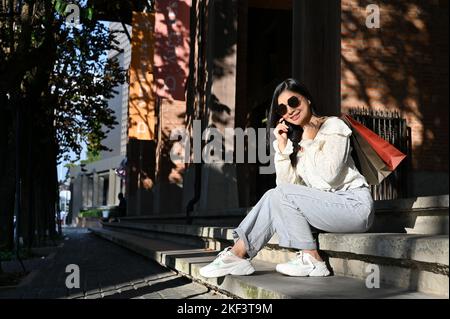 Giovane e attraente viaggiatore asiatico con occhiali da sole e borse per lo shopping seduti sulle scale della strada. Concetto di viaggio e stile di vita urbano, vista posteriore Foto Stock