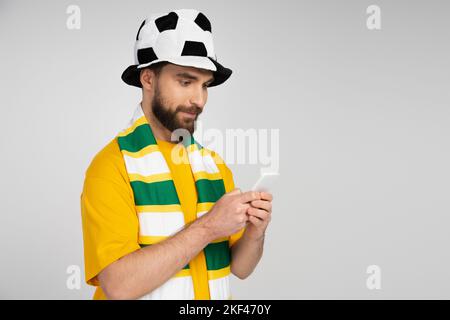 uomo concentrato in cappello tifoso di calcio e sciarpa a righe facendo scommesse sportive su smartphone isolato su grigio, immagine stock Foto Stock