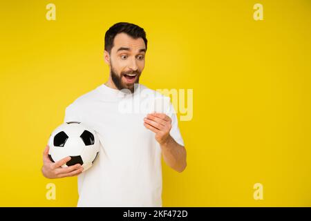 stupito e felice tifoso sportivo con palla di calcio guardare partita di calcio sul cellulare isolato sul giallo, immagine stock Foto Stock