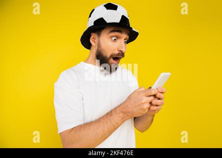 scioccato uomo in cappello tifoso di calcio guardando campionato sul cellulare isolato sul giallo, immagine stock Foto Stock