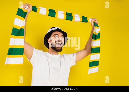tifoso sportivo overjoyed in cappello gridare mentre tenendo sciarpa a righe in mani sollevate isolato su giallo, immagine stock Foto Stock