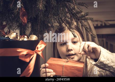 Buon Natale, buone feste. Allegro carino ragazza piccola bionda apre un regalo di Natale Foto Stock