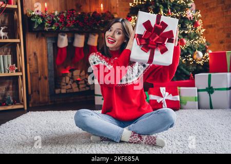 Foto di felice signora soddisfatto seduta pavimento mano tenere babbo natale presente pensare indovinare cosa all'interno pacchetto preferito casa tempo al coperto Foto Stock