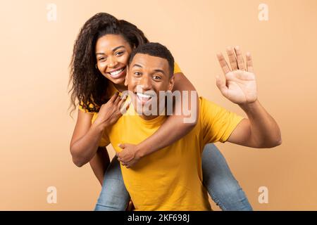 Coppia nera romantica che si diverte insieme, l'uomo che ondola alla macchina fotografica e piggybacking la sua ragazza su sfondo pesca Foto Stock