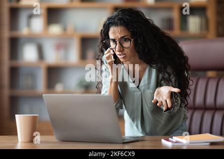 Furiosa donna d'affari mediorientale che ha una conversazione telefonica, guardando il notebook Foto Stock