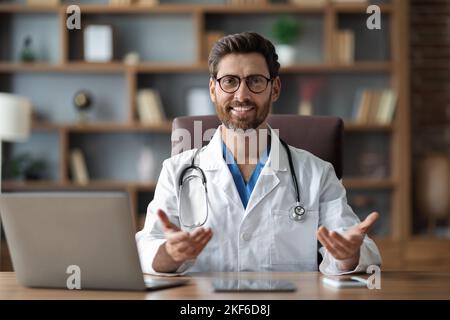 Consultazione medica. Bel medico maschio seduto alla scrivania e parlando alla macchina fotografica Foto Stock