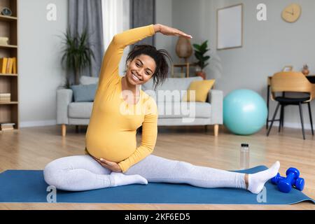 Giovane donna incinta nera sorridente con pancia grande fare esercizi, praticare yoga, allungare la mano Foto Stock