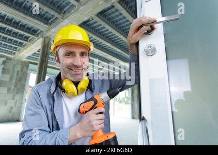 un lavoratore sorridente installa una serratura della porta utilizzando un cacciavite wireless Foto Stock