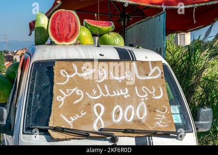 Molti cocomeri in vendita. Chiuso e mezzo aperto e accatastato su casse. Esposto di fronte a un mini-camion con una lavagna scritta a mano araba. Foto Stock