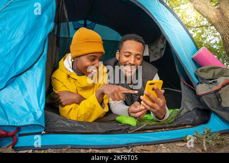 Felice coppia nera utilizzando il cellulare e seduto in tenda durante il campeggio divertirsi insieme Foto Stock