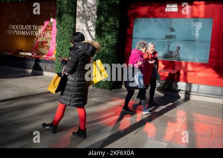 Gli amanti dello shopping passeranno attraverso la luce rossa riflessa e passeranno una vetrina natalizia all'esterno di Selfridges sotto il sole d'autunno a Oxford Street, nel West End della capitale, il 16th novembre 2022, a Londra, Inghilterra. Foto Stock