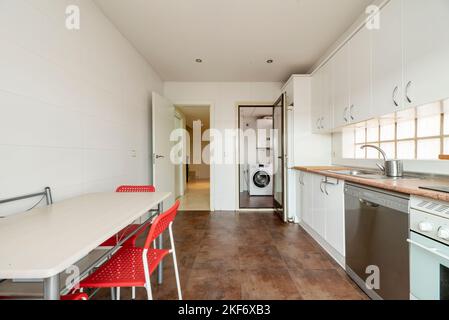 Cucina con armadi in legno bianco, elettrodomestici da incasso e cucina da pranzo con sedie rosse Foto Stock