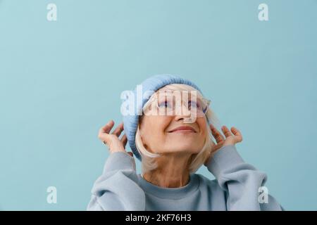 Bella donna anziana sorridente in occhiali che regola il suo cappello invernale e guarda verso l'alto su sfondo blu isolato Foto Stock