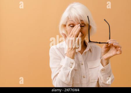 Donna anziana stanca in tuta d'affari con occhiali in mano massaggiando il suo ponte nasale su sfondo marrone isolato Foto Stock