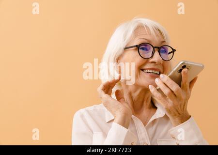 Bella donna anziana in tuta di affari e occhiali che parlano al telefono su sfondo marrone isolato Foto Stock