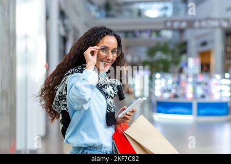 Donna d'affari shopping. Giovane donna ispanica che cammina e fa shopping in un centro commerciale. Tiene un telefono e pacchetti colorati con le merci in mano, tiene sugli occhiali, guarda la macchina fotografica e sorride. Foto Stock