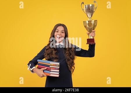 Studentessa entusiasta in divisa scolastica che celebra la vittoria con il trofeo. Premio vincente per l'adolescente su sfondo giallo. Libretto di custodia per bambini con Foto Stock