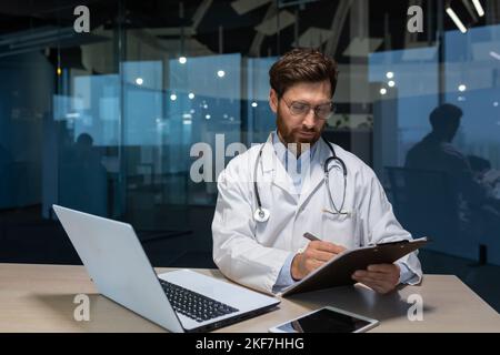 Un giovane medico maschile in un cappotto, indossando occhiali e uno stetoscopio lavora in un ufficio in un ospedale a un tavolo con un computer portatile. Scrive la storia delle cose rare, lavora con i documenti, mantiene una cartella. Foto Stock