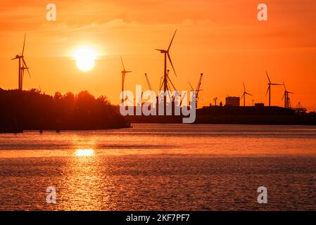 Edifici industriali con turbine eoliche e gru portuali su un porto fluviale che si affaccia su un cielo fiero al tramonto Foto Stock