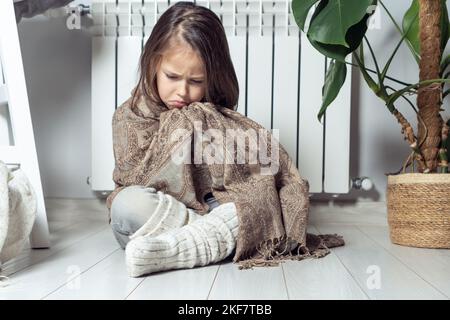 Depressa, piangeva, ragazza piccola seria avvolta in plaid caldo, provando a scaldarsi, mantenere caldo con vestiti di lana, sedersi sul pavimento vicino al radiatore di riscaldamento Foto Stock