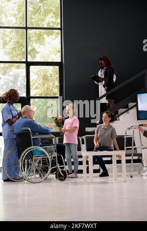 Nonna che dà bouquet di fiori al nonno disabile durante il check up visita appuntamento in ospedale sala d'attesa. Paziente in sedia a rotelle che discute il trattamento farmacologico con l'infermiere medico Foto Stock