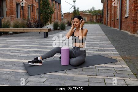 Atleta femminile seduto sul tappetino sportivo durante gli allenamenti all'aperto e che si diverte ad ascoltare le canzoni preferite con le cuffie Foto Stock